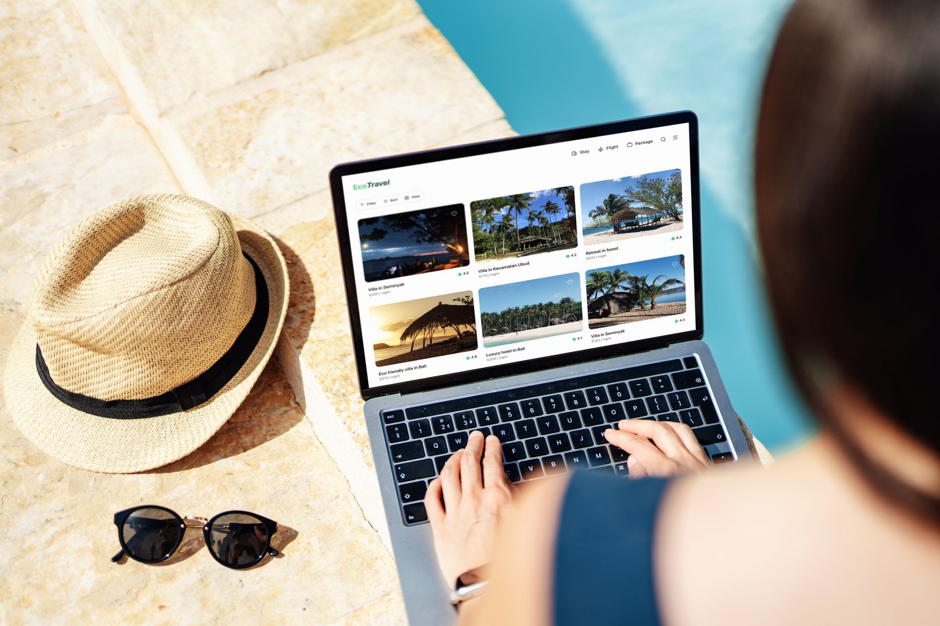 Person using laptop poolside, browsing tropical vacation images; hat and sunglasses nearby.