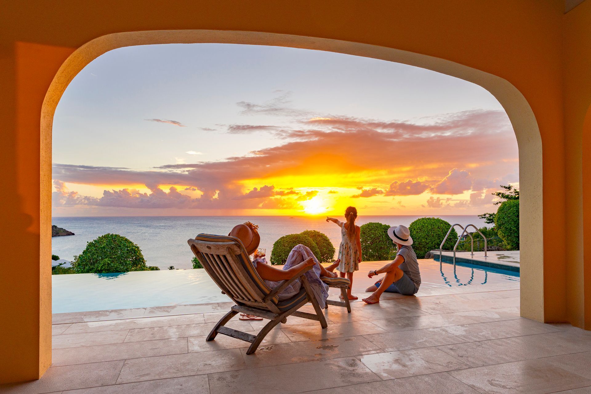 Sunset view through an archway, three people by a pool, ocean horizon.