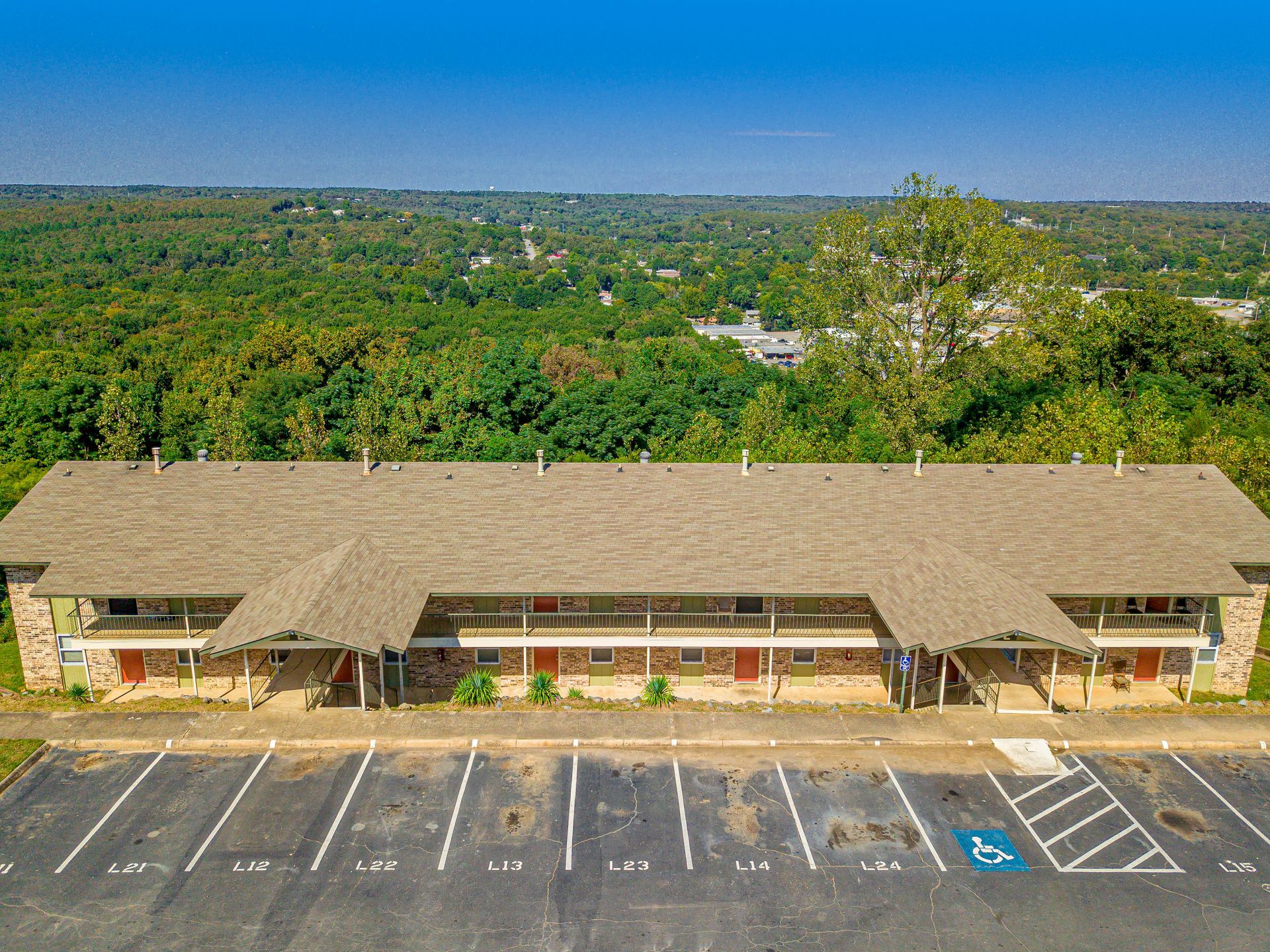 Two-story beige apartment building with parking, set against a backdrop of trees and a clear sky.