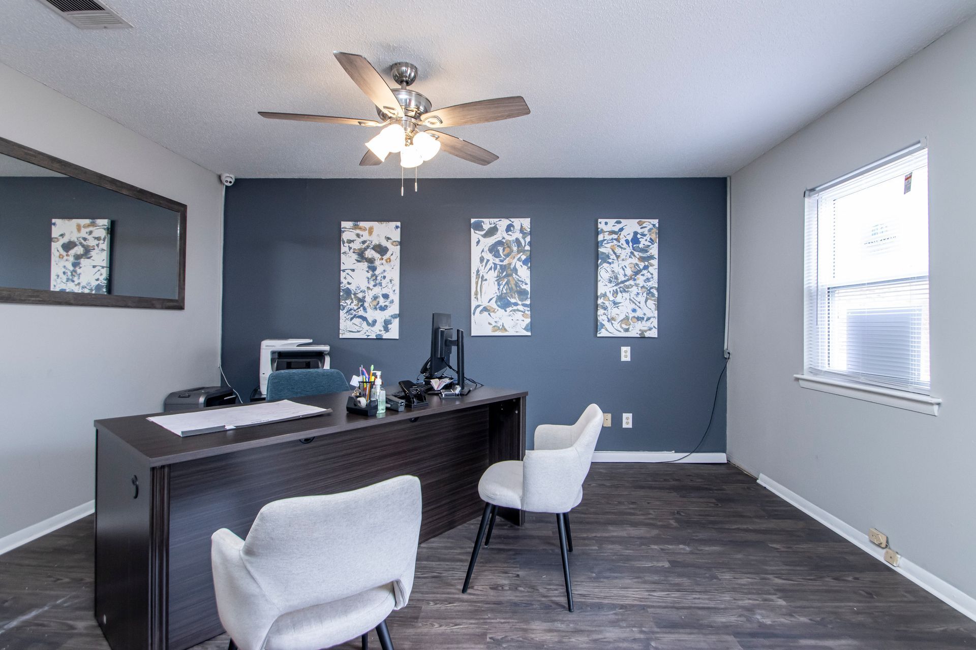 Modern office with desk, chairs, artwork on blue wall, and ceiling fan.