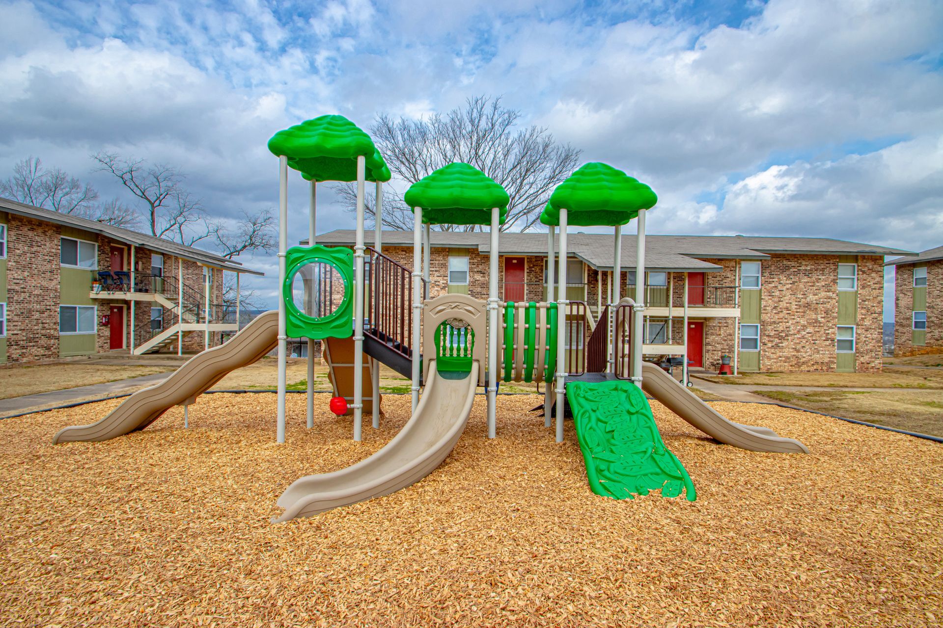 Playground with slides and green structures, set against apartment buildings and a cloudy sky.