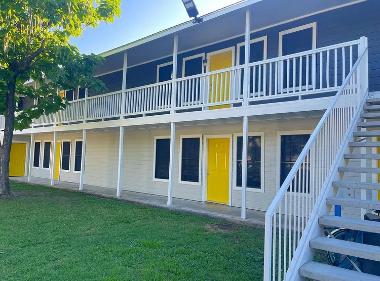 Two-story building with tan siding, white railings, yellow doors, and a grassy lawn.