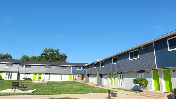 Apartment complex exterior with green grass, blue and grey buildings under a clear blue sky.