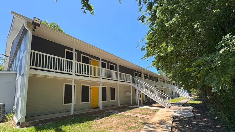 Two-story apartment building with white balconies, yellow doors, and outdoor staircases; sunny day.