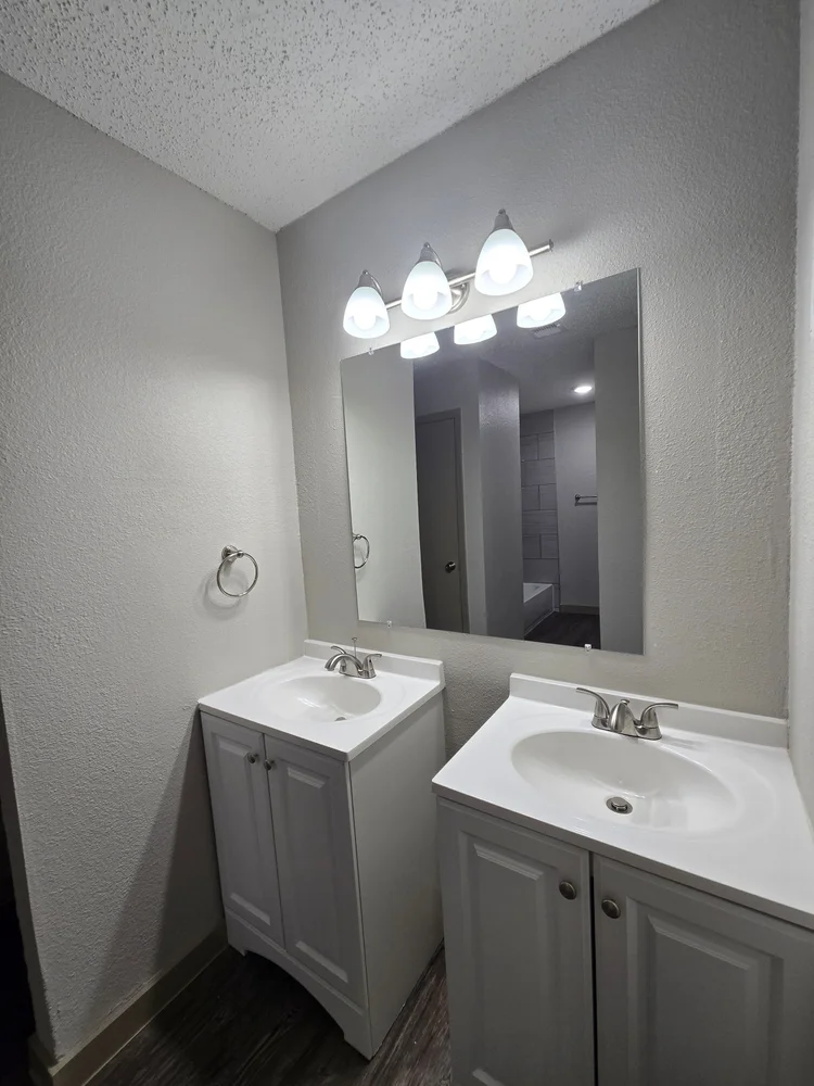 Bathroom with two white vanities, a mirror, and a three-light fixture. Gray walls and dark flooring.