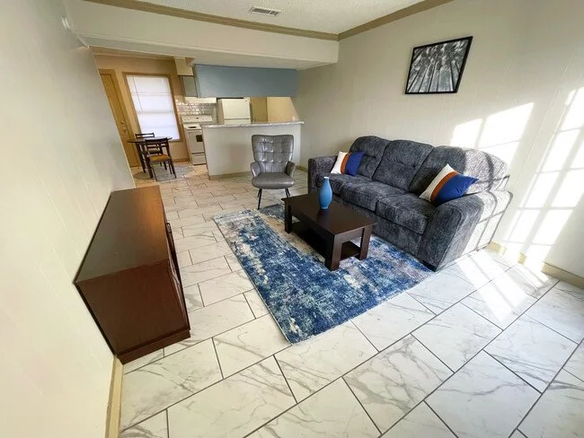 Living room with blue sofa, rug, and TV stand; kitchen in the background.