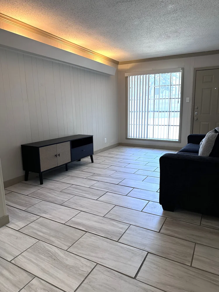 Living room with gray tiled floor, black TV stand, and dark sofa near a window and door.