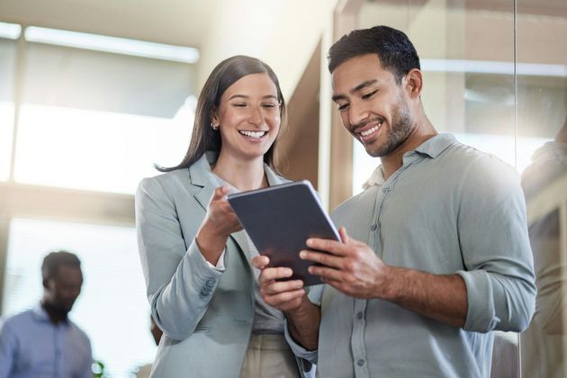 Two smiling colleagues in a brightly lit office look together at a digital tablet held by the person on the right.
