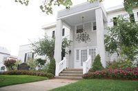 White building with columns, steps, and chandelier. Green lawn and shrubbery.