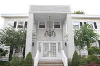 White house facade with ornate entrance, double doors, chandelier, and small front yard with shrubs.