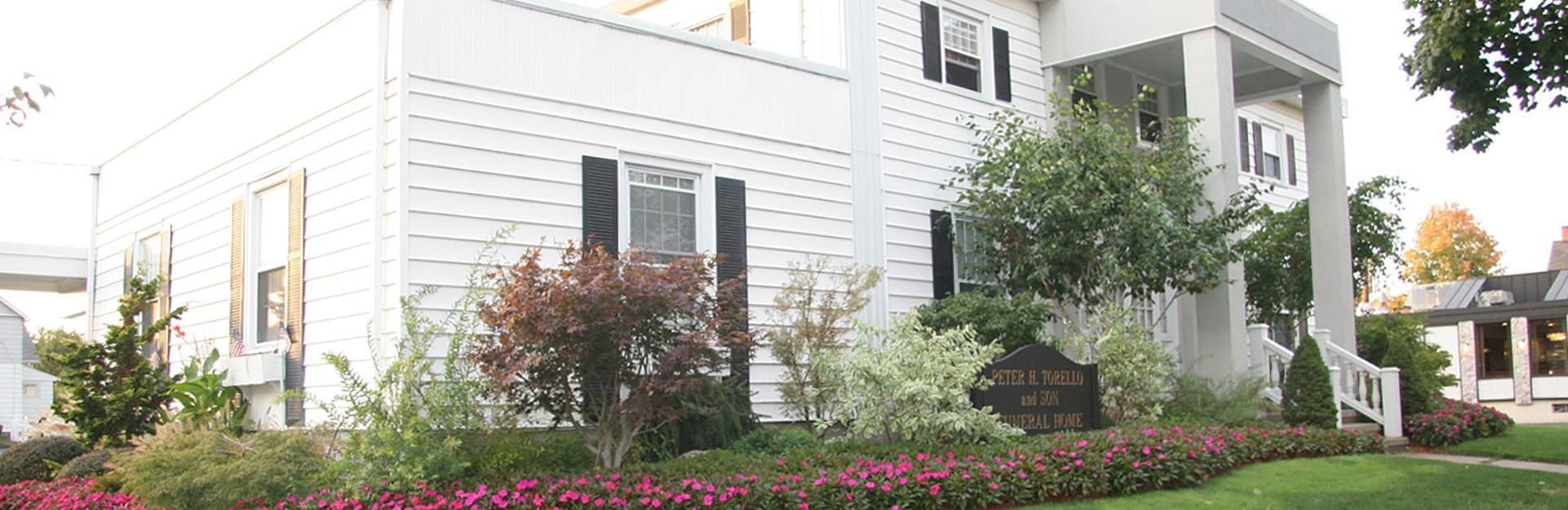 White building with black shutters and a sign surrounded by landscaping.