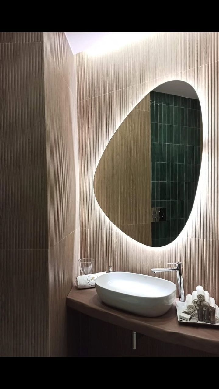 Bagno moderno con specchio ovale illuminato da una luce calda, lavandino bianco e ripiano in legno scuro.