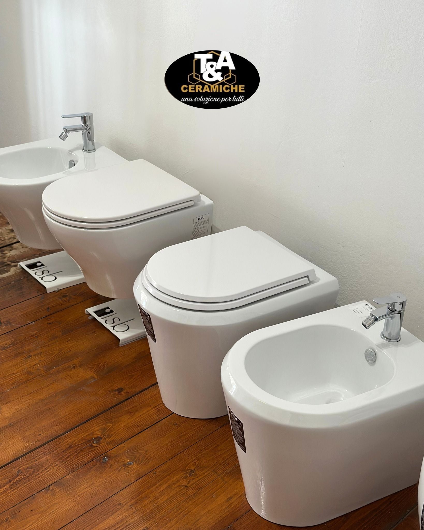 Sanitari bianchi (WC e bidet) esposti su un pavimento in legno contro una parete bianca; esposizione dei prodotti.