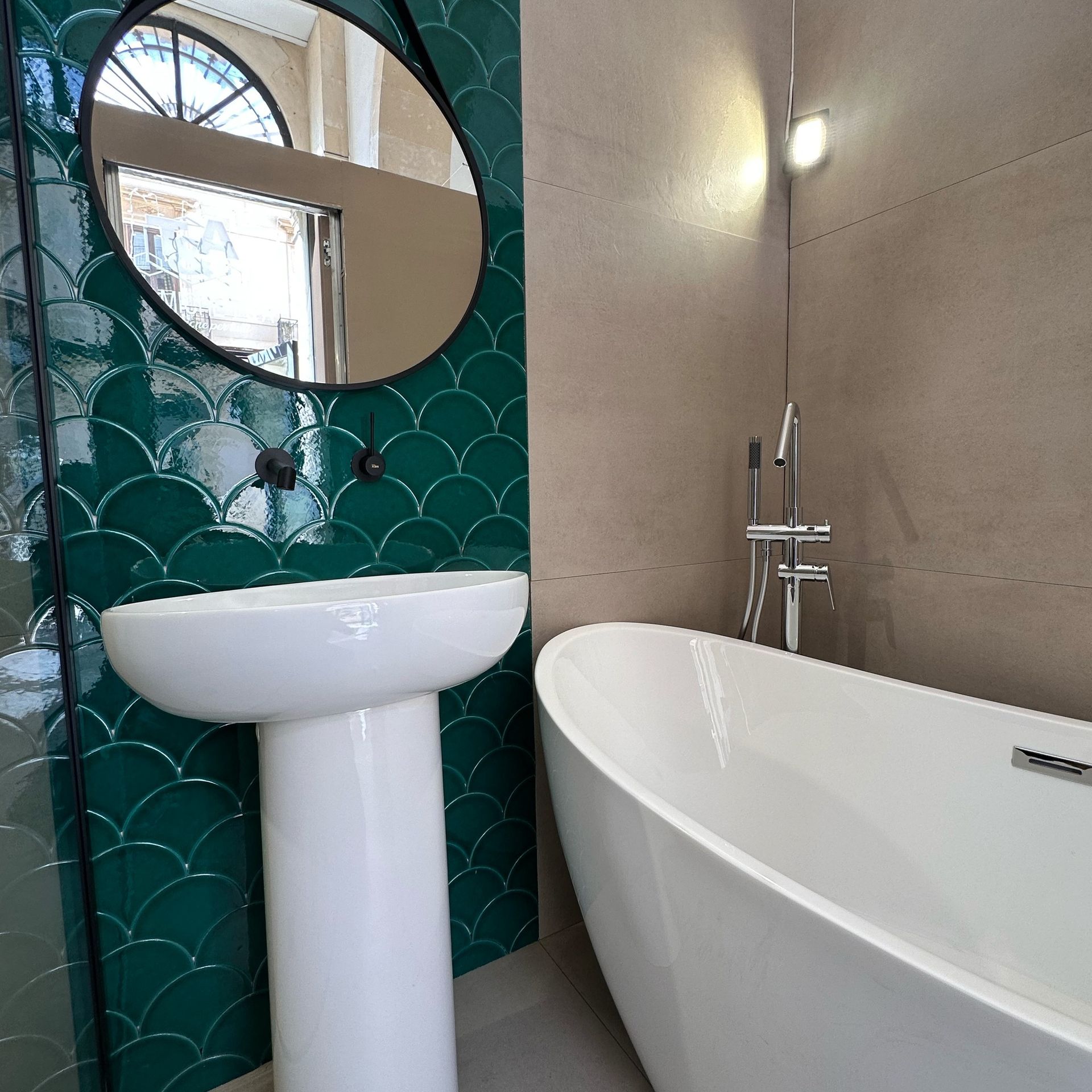 Bagno con piastrelle smerlate verde smeraldo, specchio nero rotondo, lavabo bianco con piedistallo e vasca bianca.