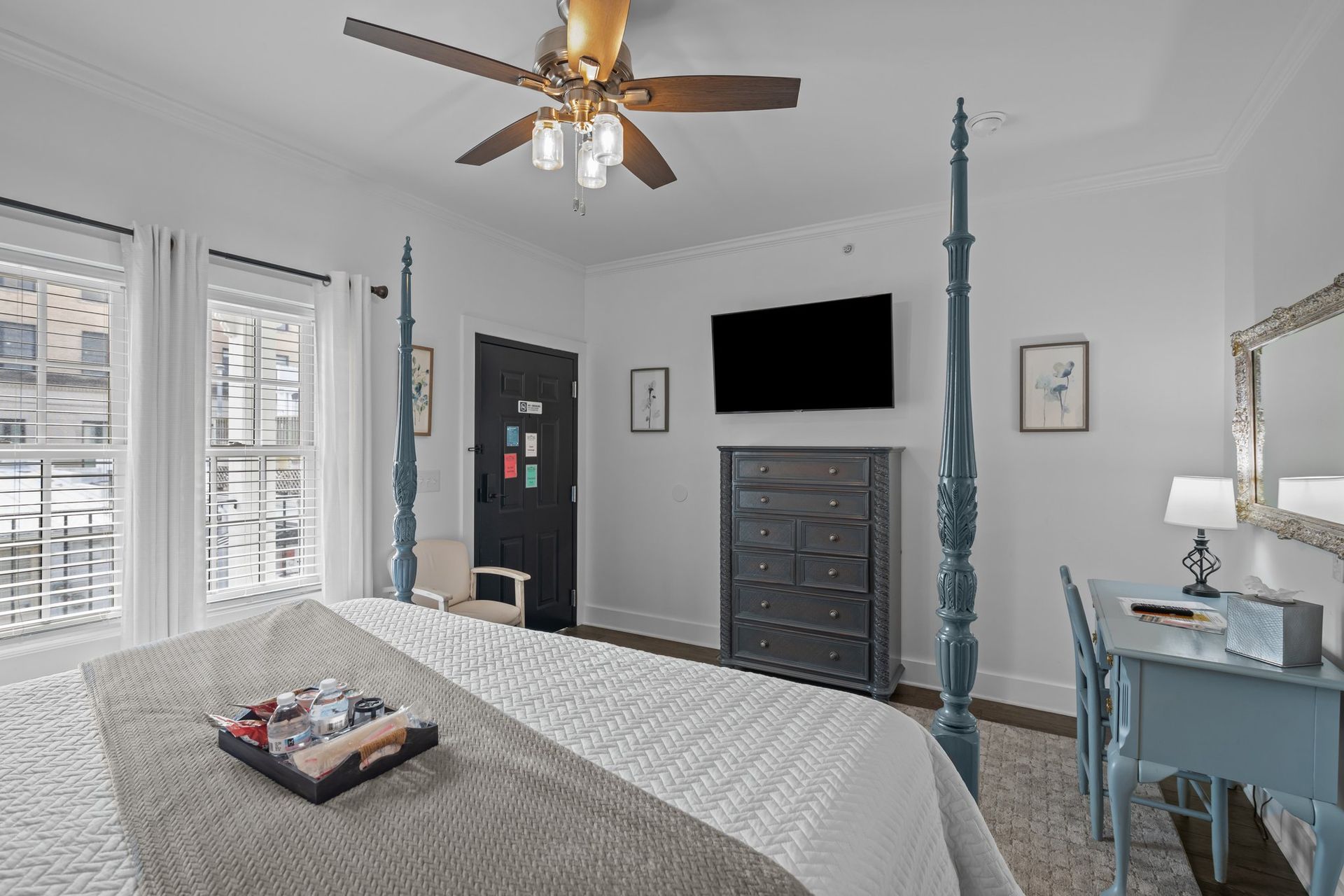 A bedroom with a four poster bed , dresser , desk , television and ceiling fan.