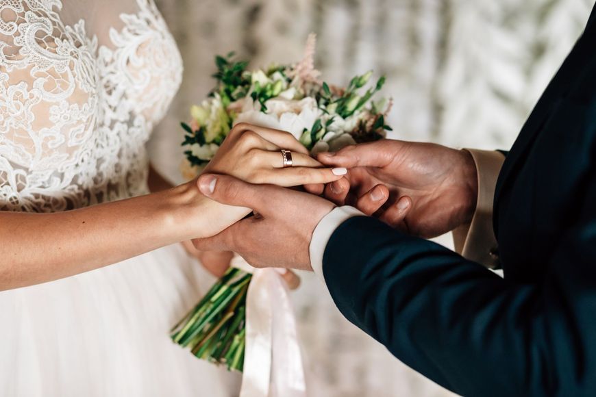A bride and groom are holding hands and the groom is putting a wedding ring on the bride 's finger.