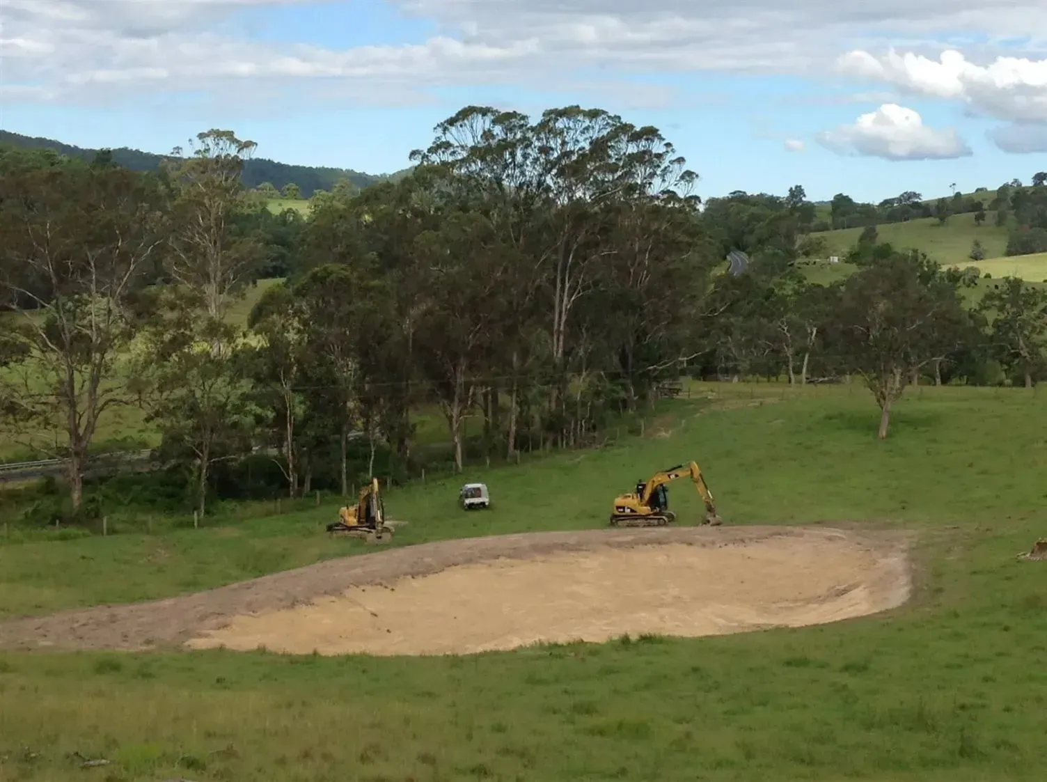 A Yellow Excavator is Working in a Grassy Field With Green Horizons — S M & A J Gilbert Earthmoving in Mitchells Island, NSW
