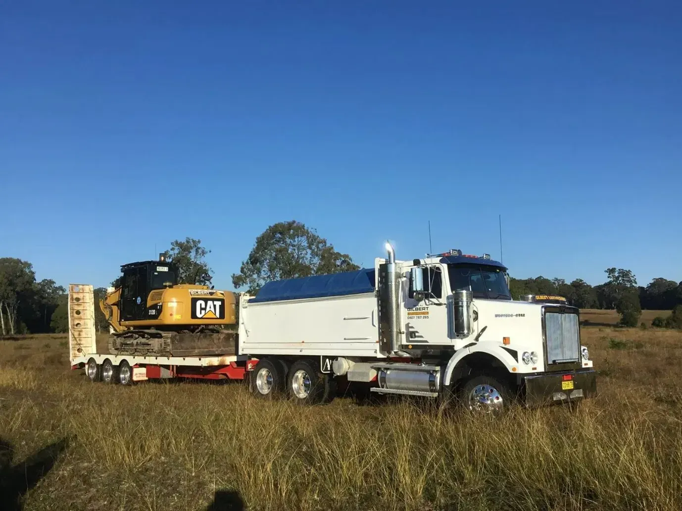 A Dump Truck With Orange Cat Excavator on the Back is Parked in a Field — S M & A J Gilbert Earthmoving in Mitchells Island, NSW