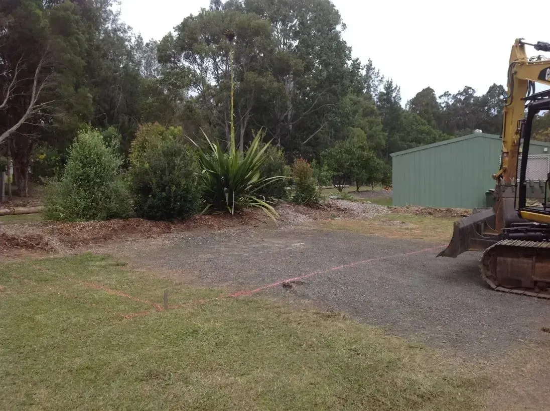 A Yellow Excavator is Driving Down a Gravel Road in a Yard With Trees — S M & A J Gilbert Earthmoving in Mitchells Island, NSW