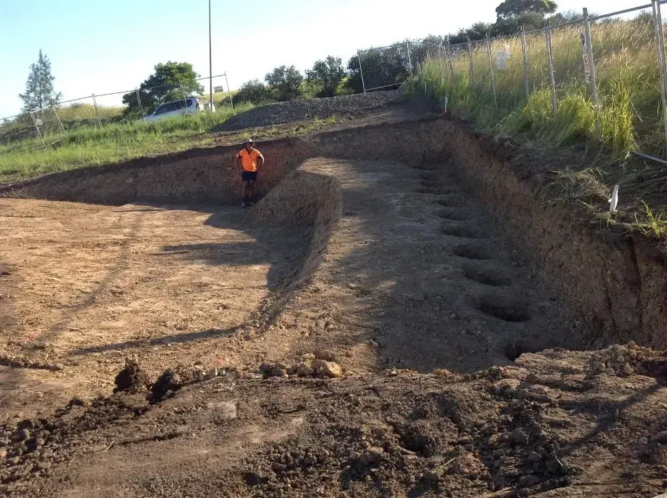 A Man in an Orange Shirt is Standing in a Dirt Field — S M & A J Gilbert Earthmoving in Mitchells Island, NSW