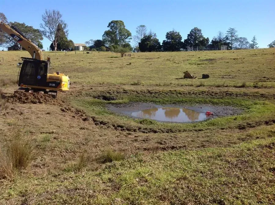 A Yellow Excavator is Digging a Hole in a Field Near A Puddle — S M & A J Gilbert Earthmoving in Mitchells Island, NSW
