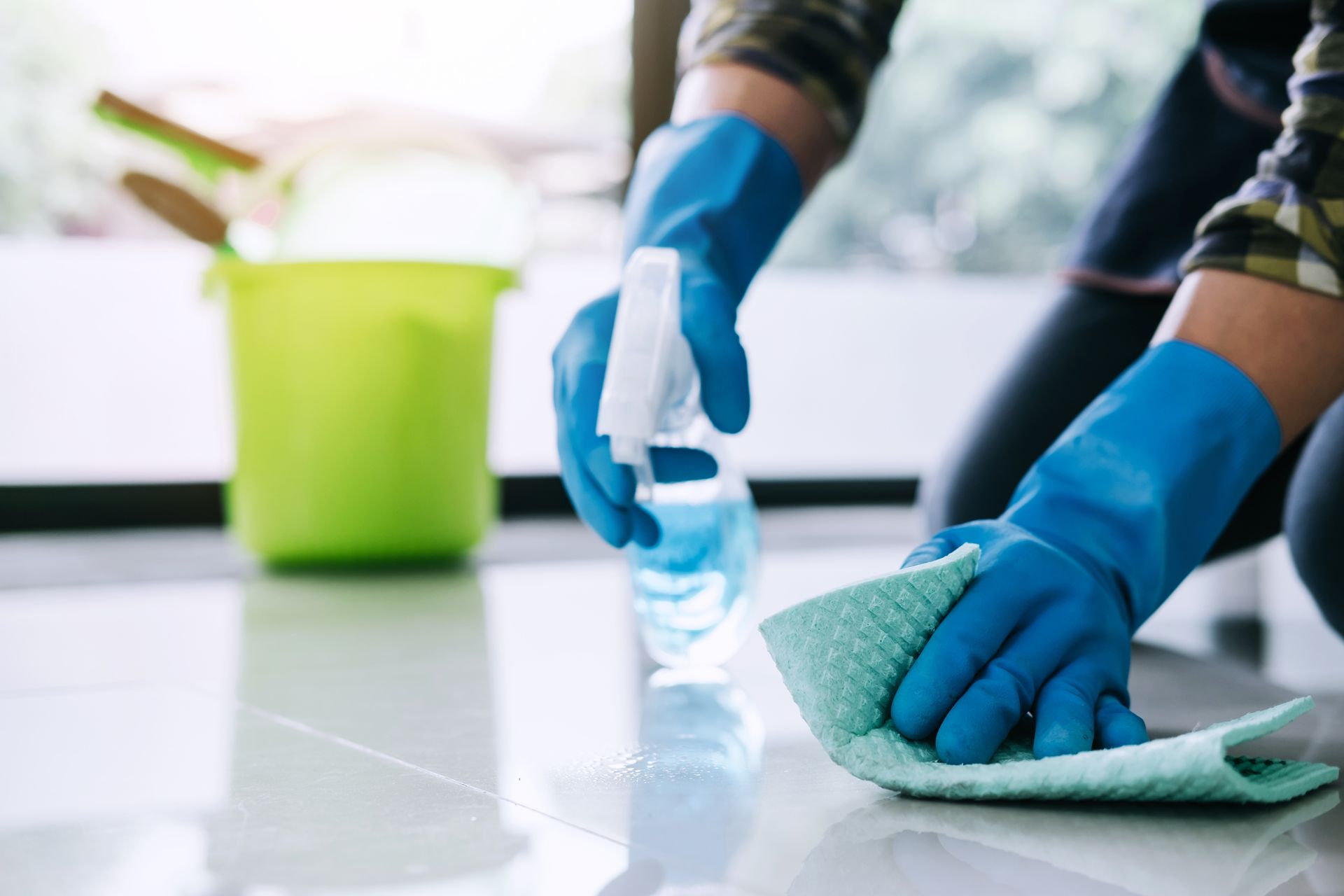 Blue-gloved hands wipe a countertop with a cloth and spray cleaner; green bucket in the background