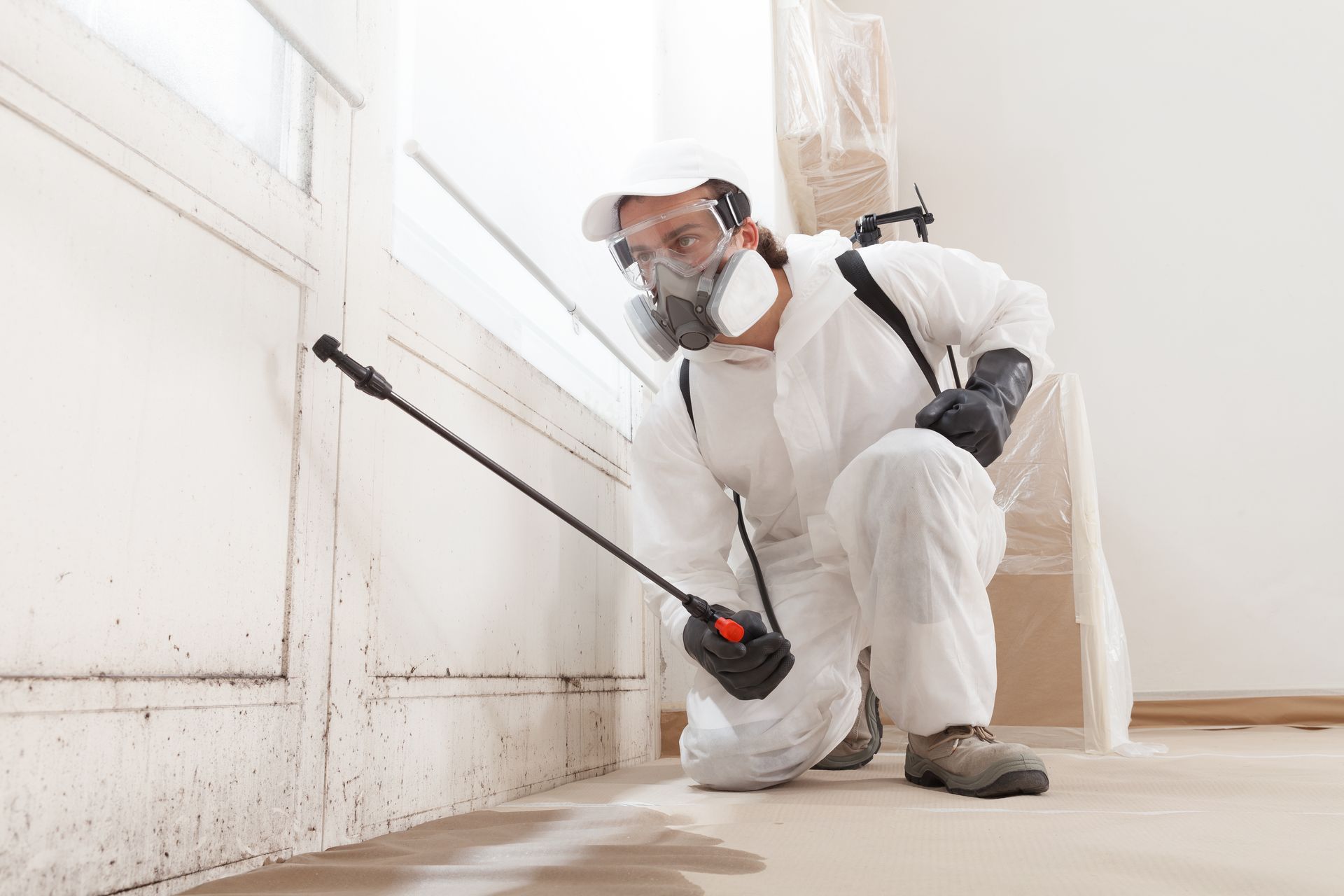 Worker in white protective gear spraying pesticides along a wall indoors