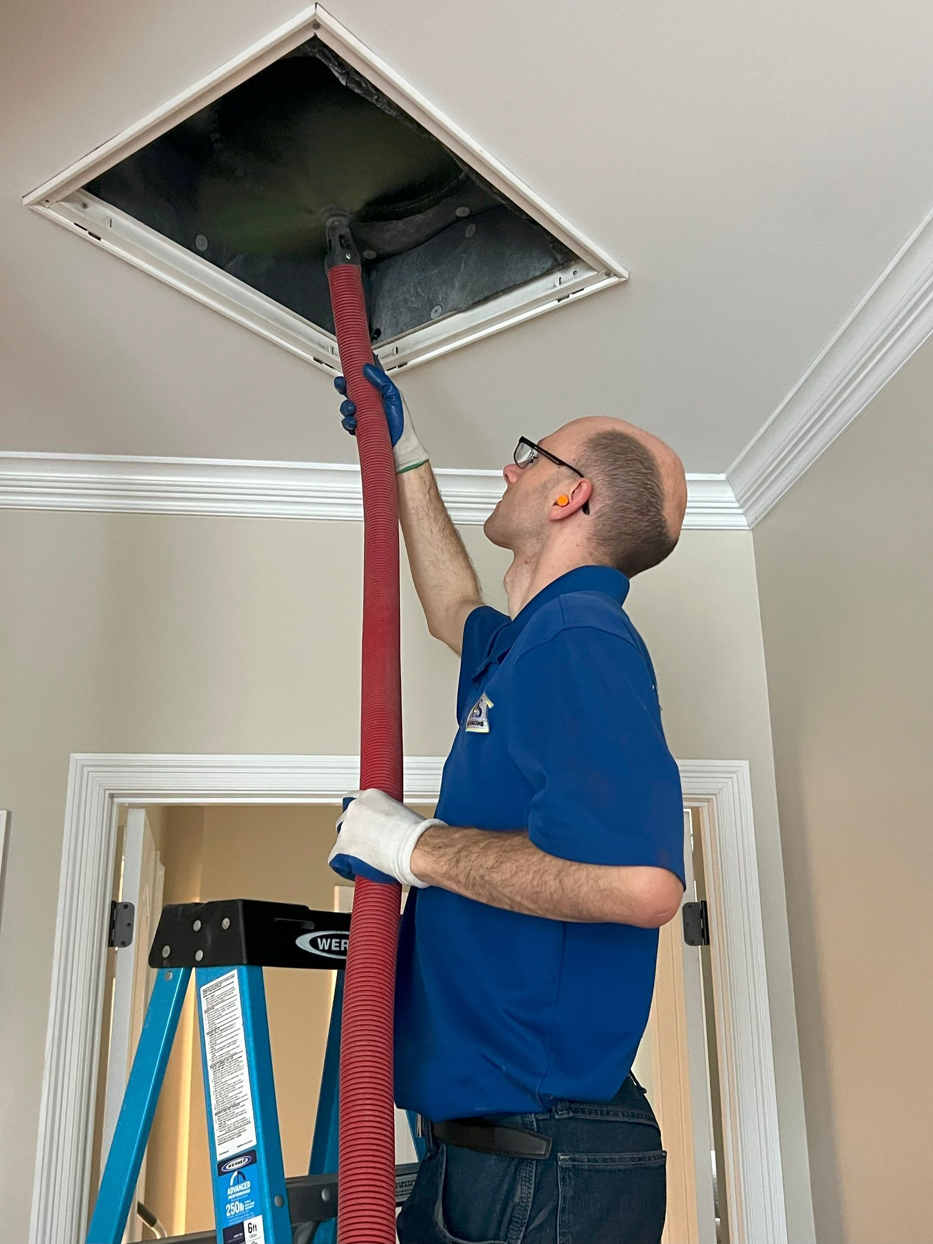 Worker using a red hose to clean an air duct opening in a ceiling room.