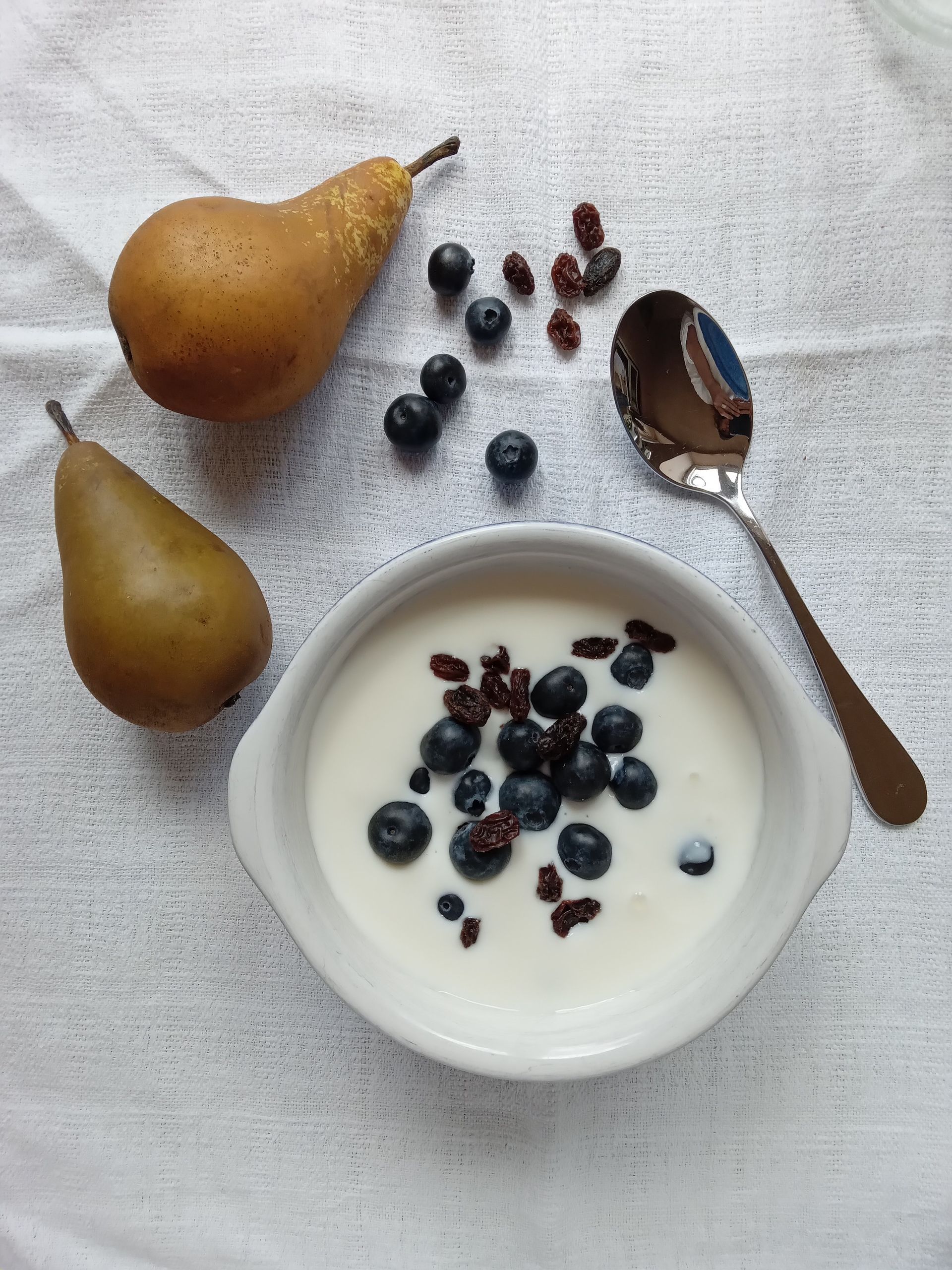 Pear and blueberry yogurt bowl with spoon on a white cloth, with scattered berries nearby