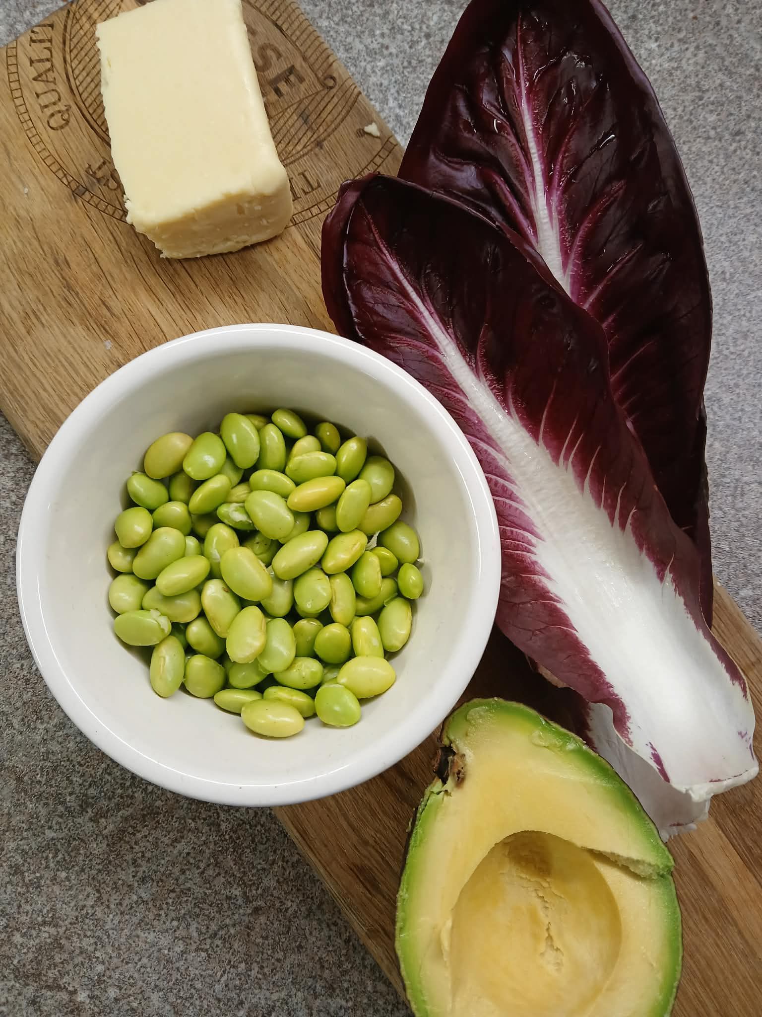 A bowl of edamame, avocado half, radicchio leaf, and butter on a textured surface.