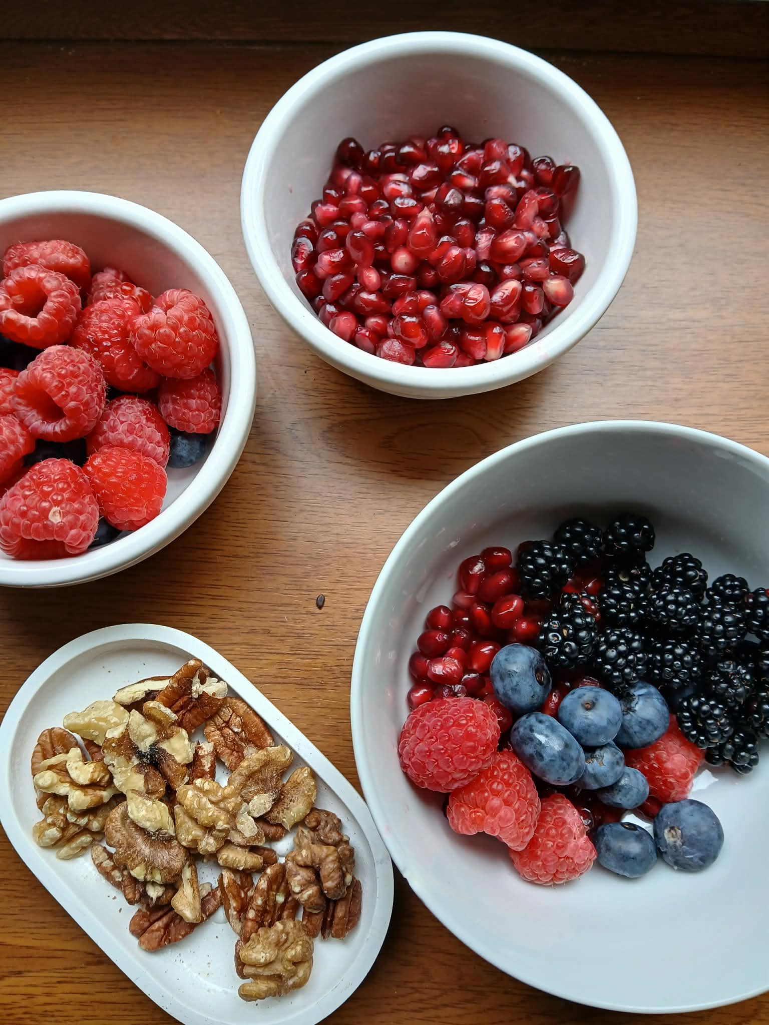 Bowls of raspberries, pomegranate seeds, mixed berries, and walnuts on a wooden table