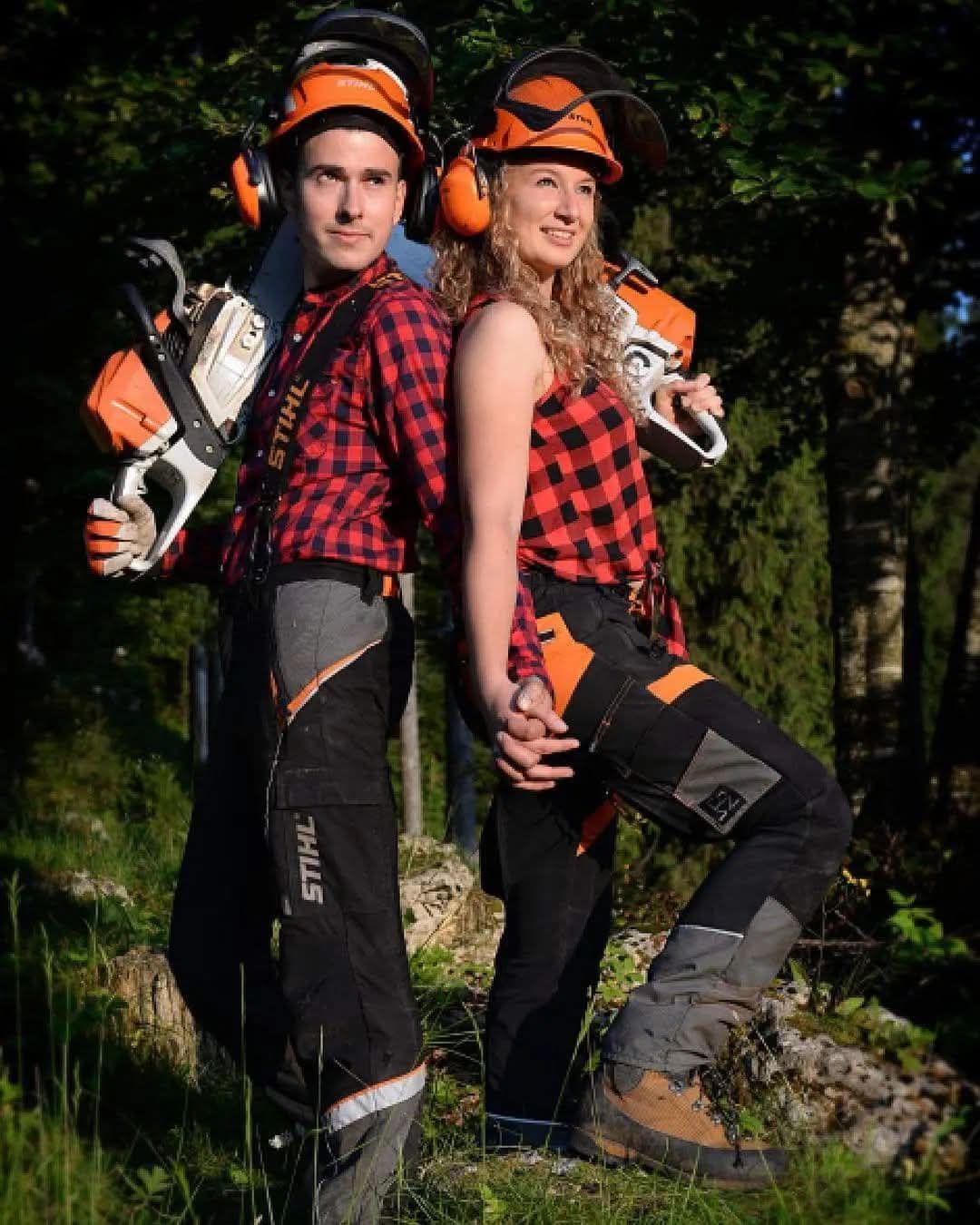 Due persone vestite da boscaioli, con in mano delle motoseghe, sono in piedi, schiena contro schiena, all'aperto.