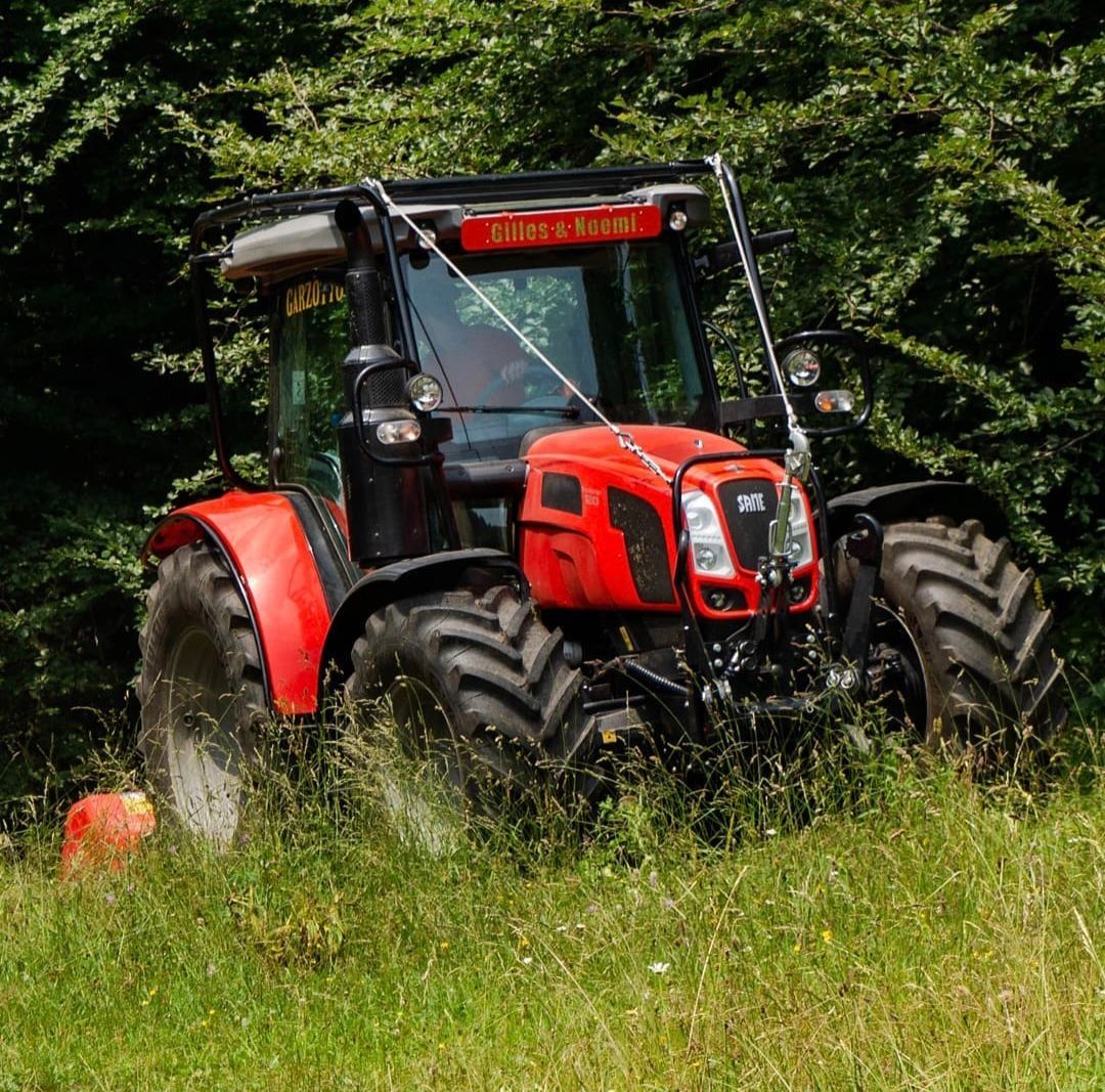 Trattore rosso e nero nell'erba alta, accanto a una siepe verde, alla luce del giorno.