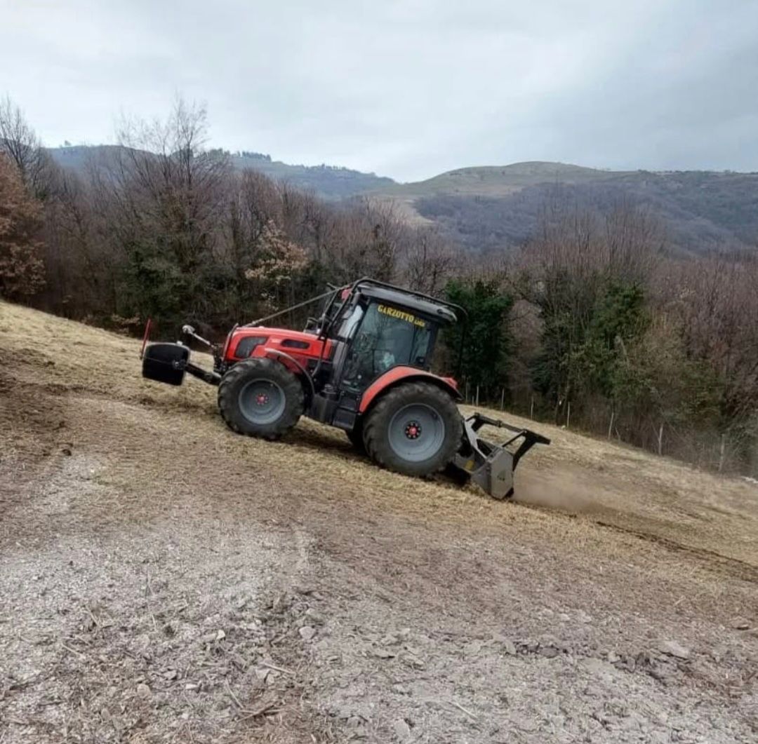 Trattore rosso al lavoro su un pendio, impegnato a falciare o arare il terreno, con uno sfondo collinare e un cielo nuvoloso.