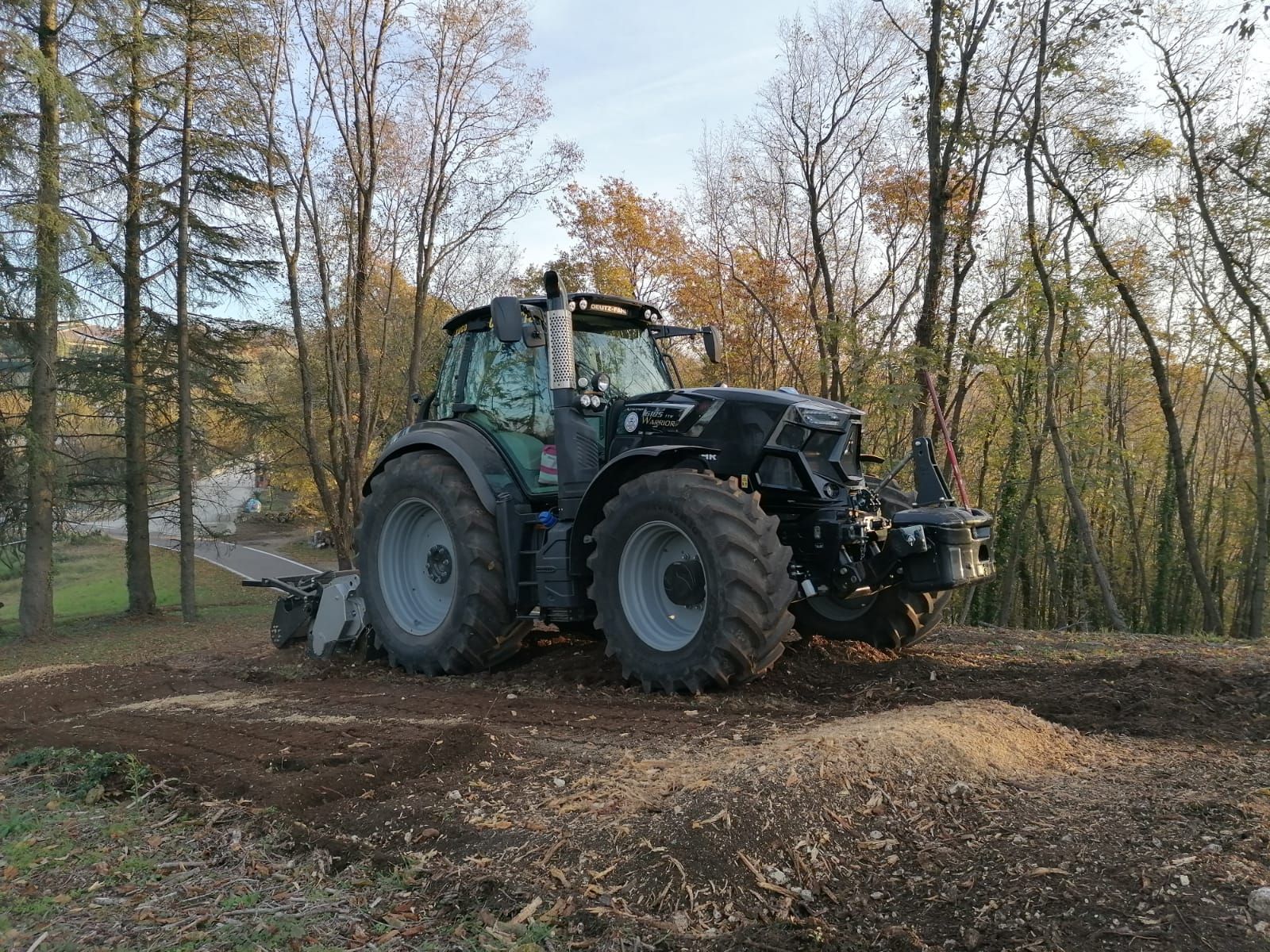 Trattore scuro che ara il terreno in una zona boscosa, terra marrone con trucioli di legno, alberi autunnali.