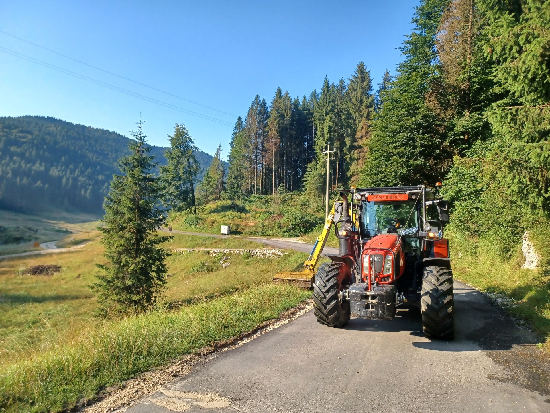 Trattore parcheggiato sulla strada accanto alla foresta. Alberi e montagne sullo sfondo sotto il cielo azzurro.