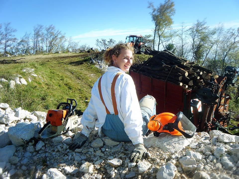 Una donna in abiti da lavoro sorride, seduta su delle rocce con una motosega e un casco protettivo. Sullo sfondo, legna e un trattore.