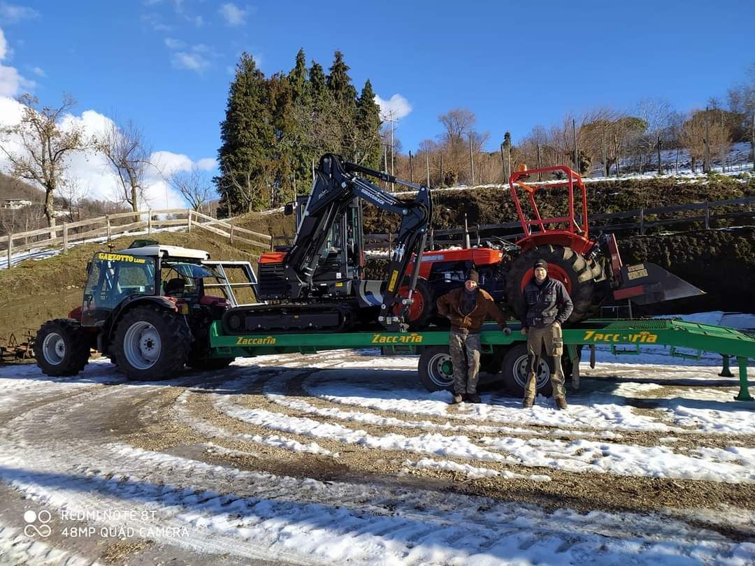 Due uomini sono in piedi davanti a un rimorchio carico di trattori e di un escavatore su una strada innevata, sotto un cielo luminoso.