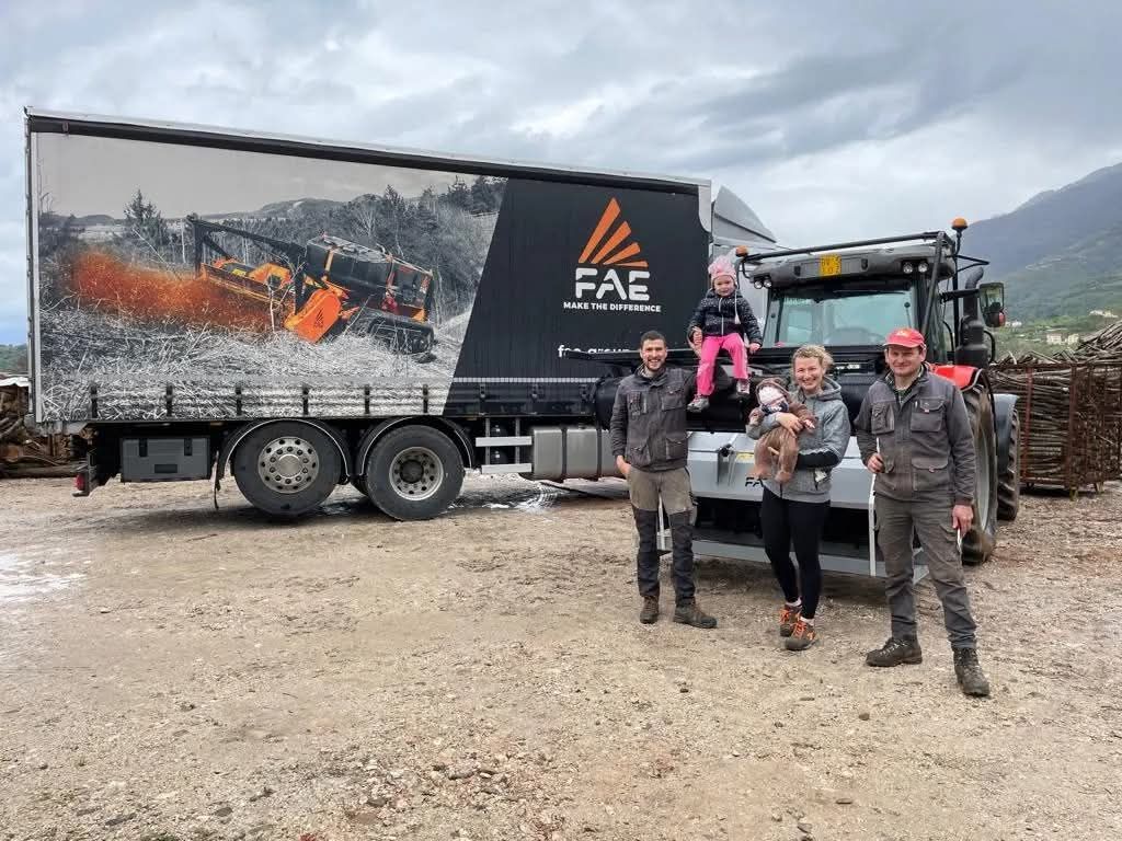 Famiglia con cane in posa davanti a un camion con il logo FAE e la grafica di un macchinario, all'esterno in un parcheggio ghiaioso.
