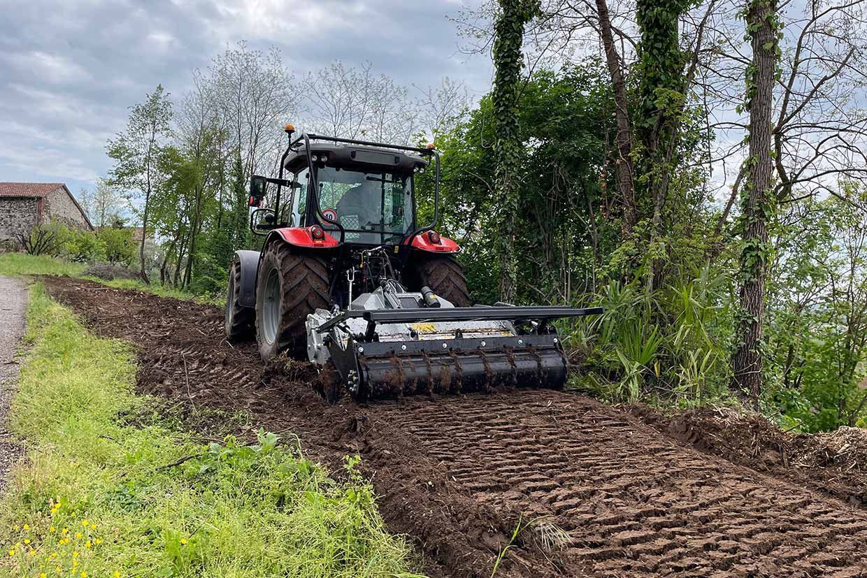 Trattore con fresa rotativa che ara un campo fangoso vicino a un'area erbosa e ad alberi.