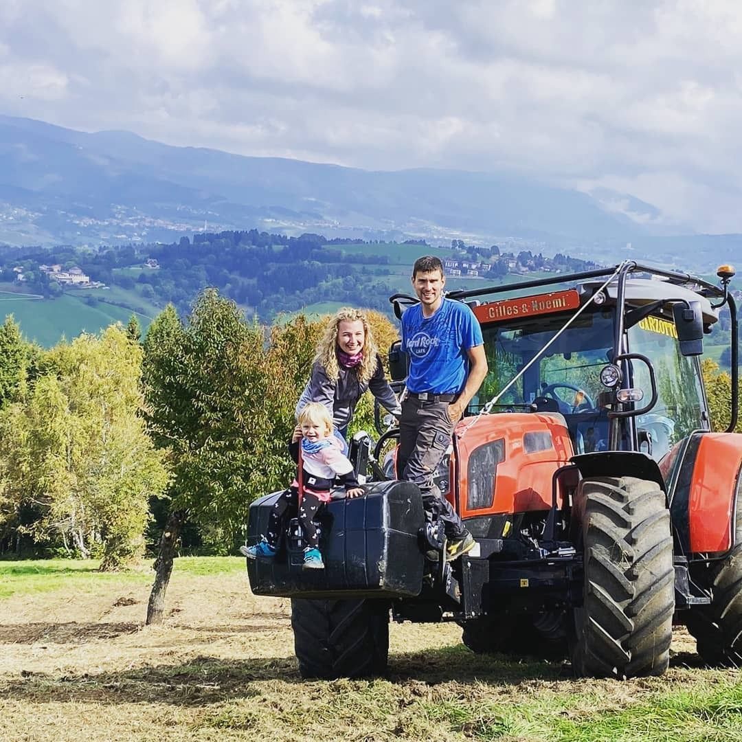 Coppia su un trattore arancione in un campo con montagne sullo sfondo.