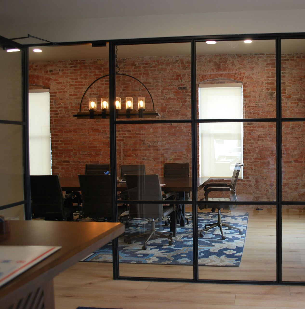 Glass-walled conference room with exposed brick, wooden table, chairs, blue patterned rug, and chandelier.