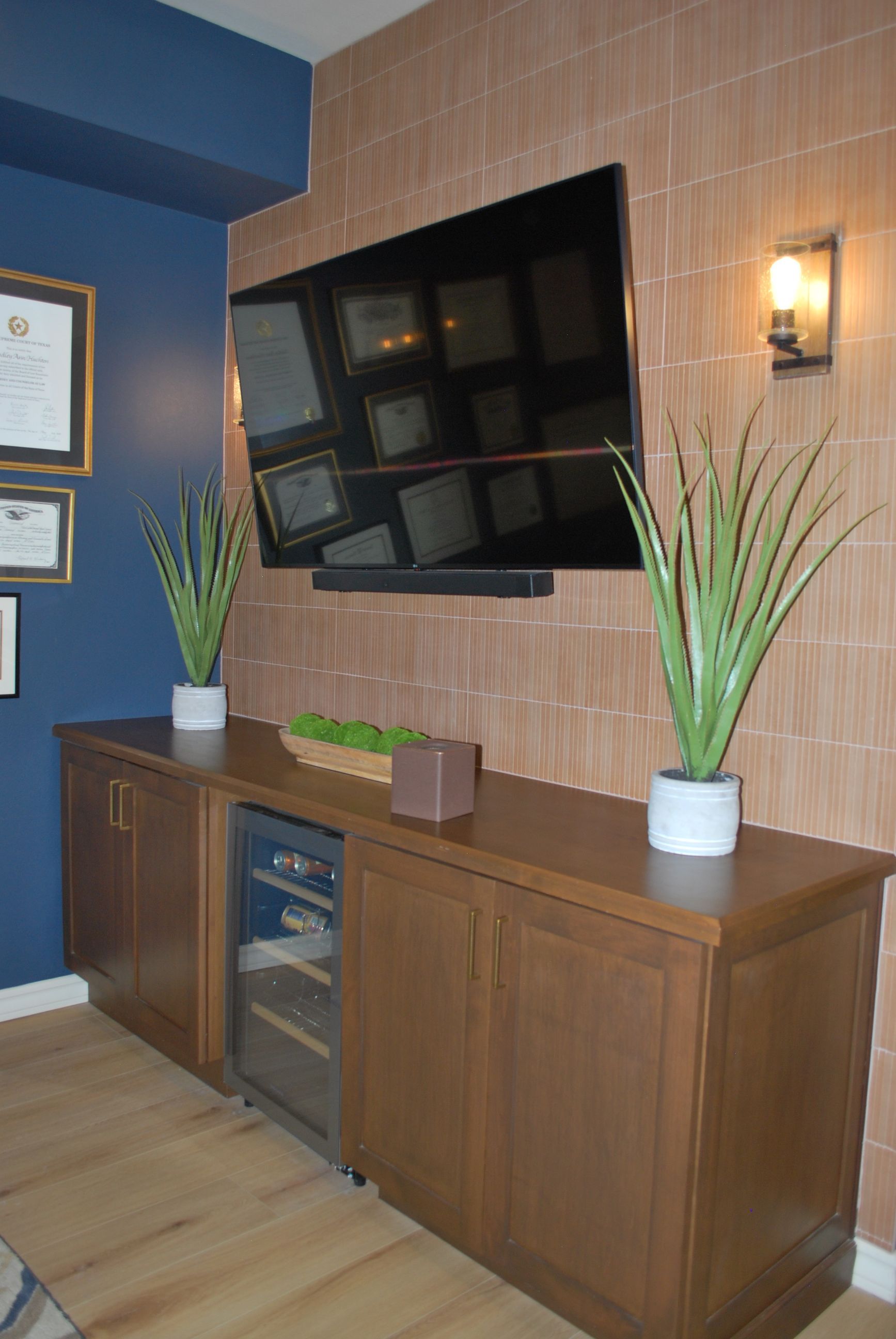 A media console with TV, wine fridge, plants, and wall sconces against bamboo wallpaper and blue wall.