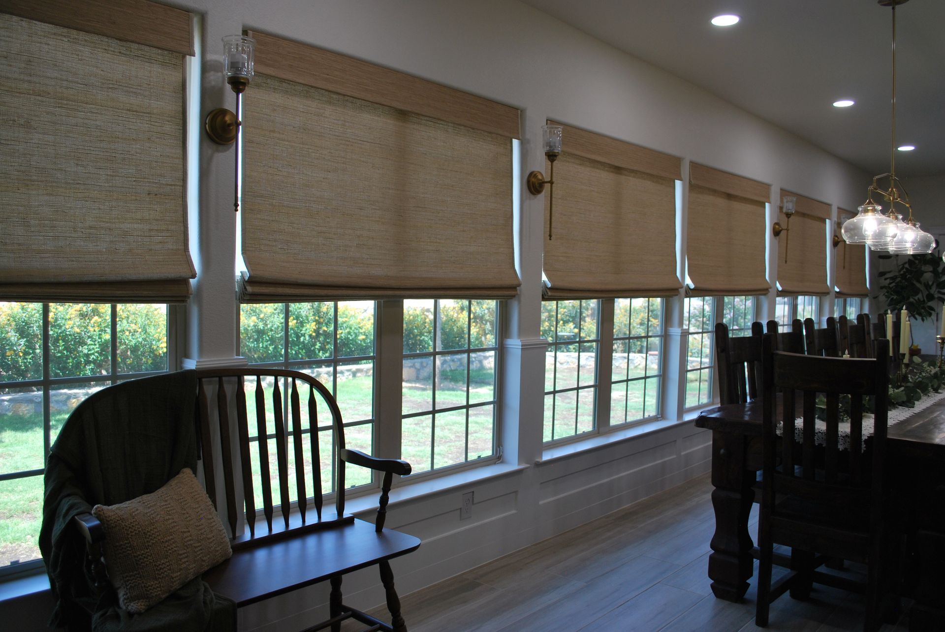Row of windows with tan woven shades, overlooking a green yard. A wooden bench and table are in view.
