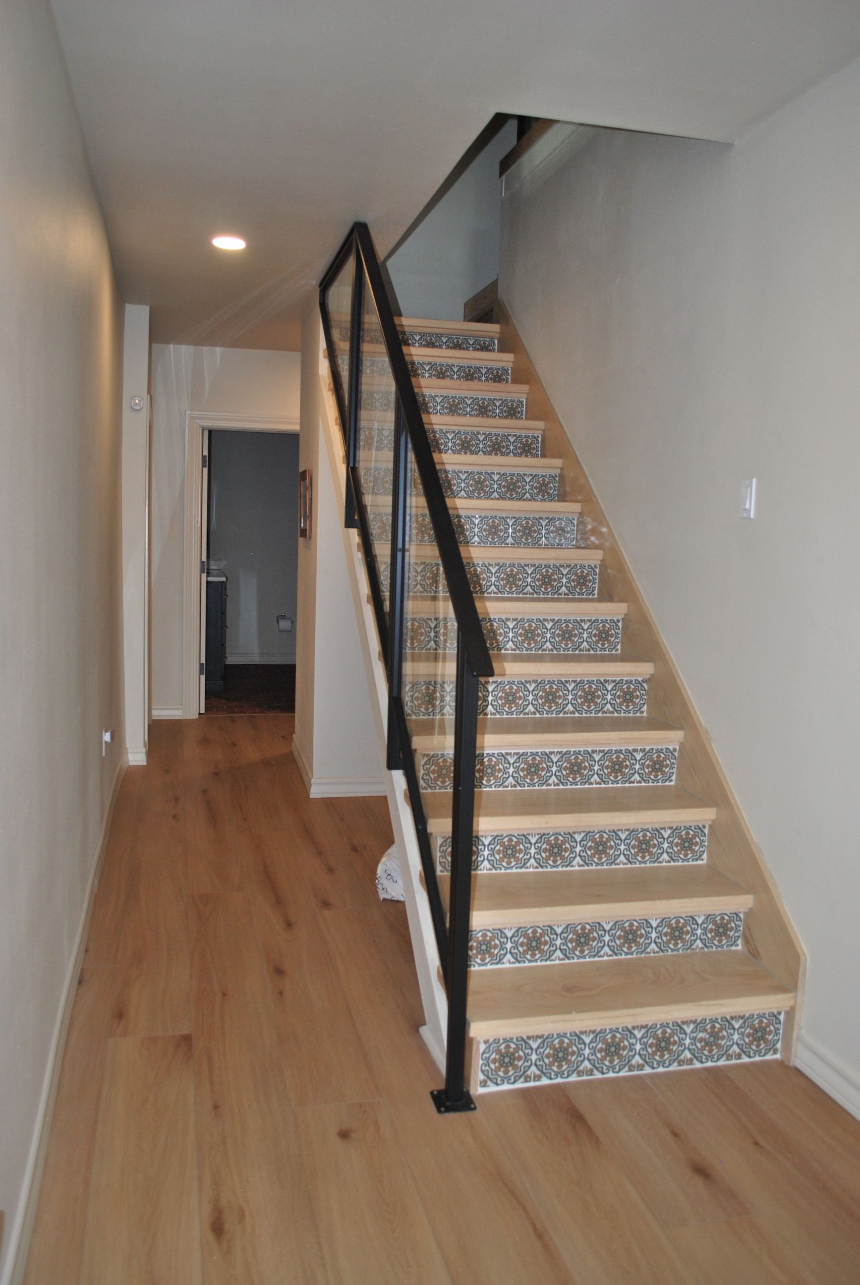 Wooden staircase with decorative tile risers, black metal handrail, light wood floor and off-white walls.