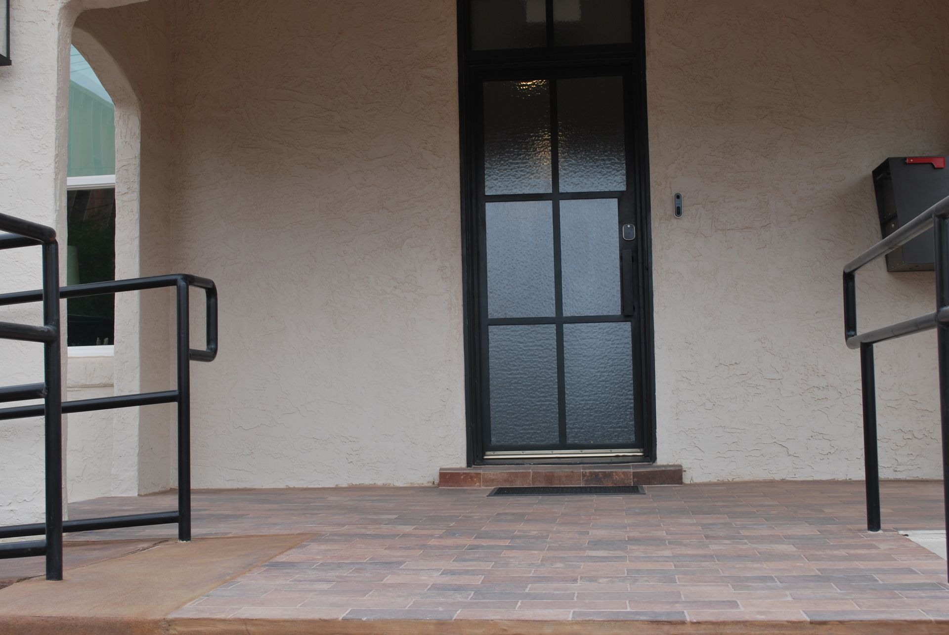A black front door with glass panels on a building's porch, accessible by a ramp with black handrails.