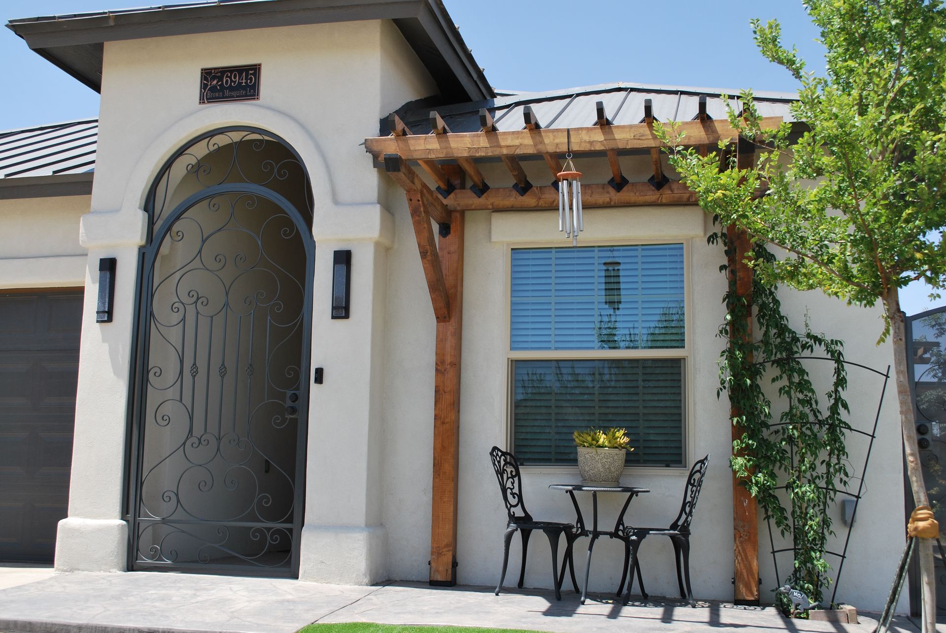 Beige stucco house with black iron gate, wooden pergola over window with small table and chairs.