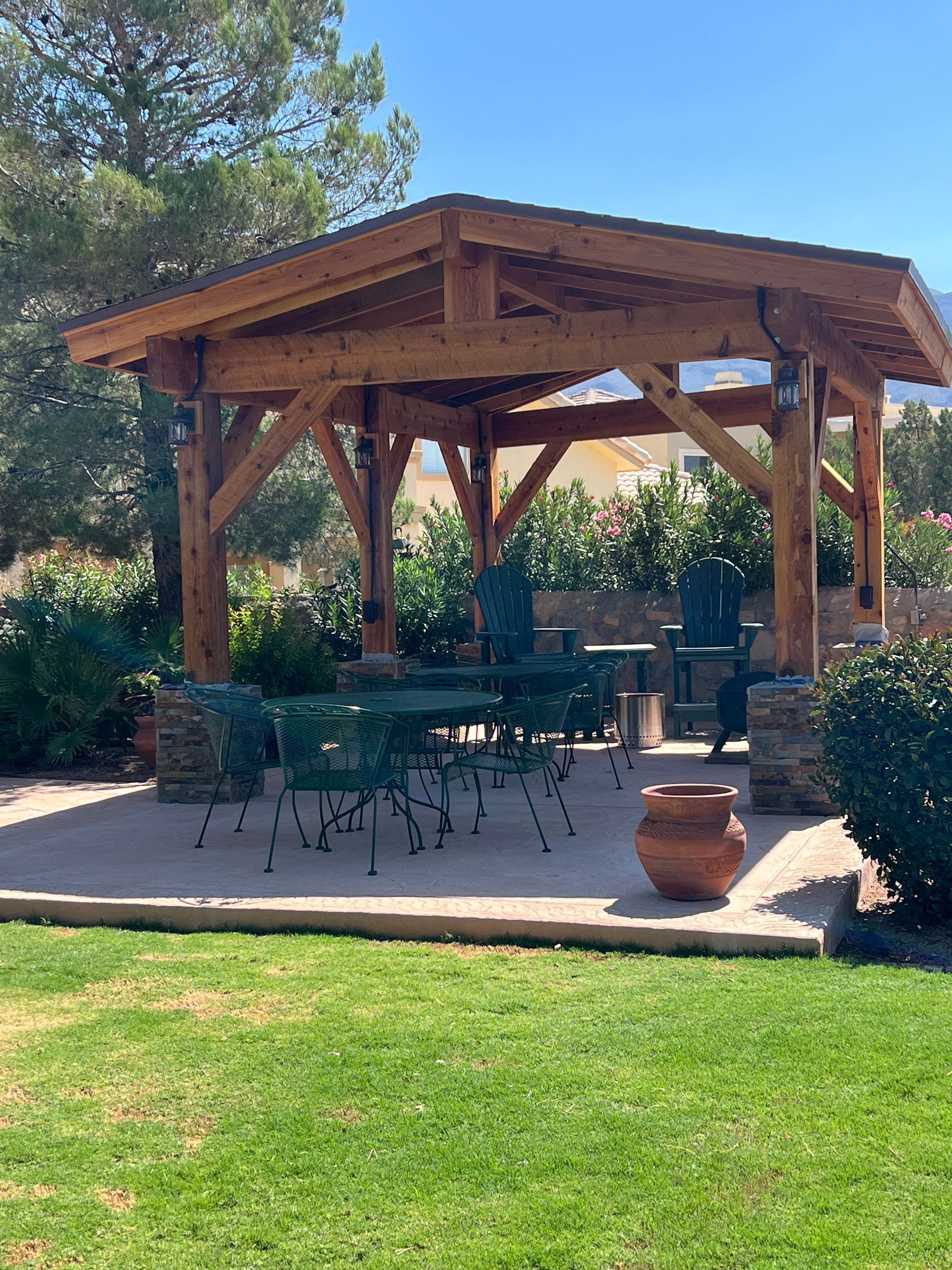 Wooden gazebo with green metal chairs and table on a patio. Overlooking garden on sunny day.