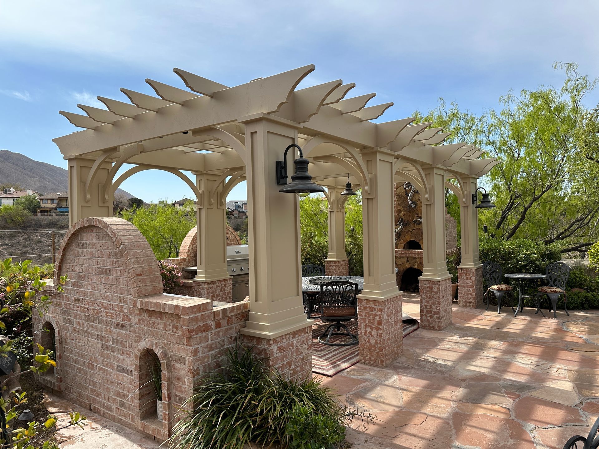 Stone outdoor fireplace under a wood-beam patio roof, with chairs and potted plants.