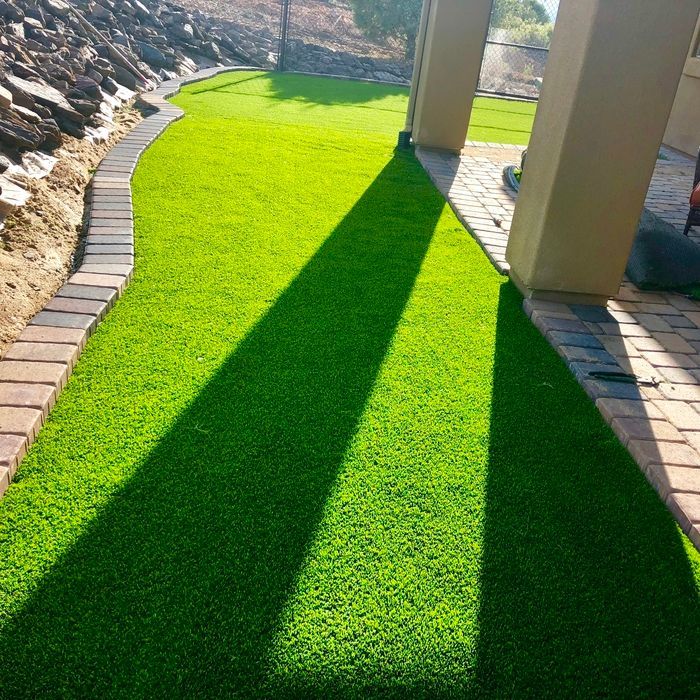 Green artificial turf pathway bordered by brick, with strong sunlight creating shadow.
